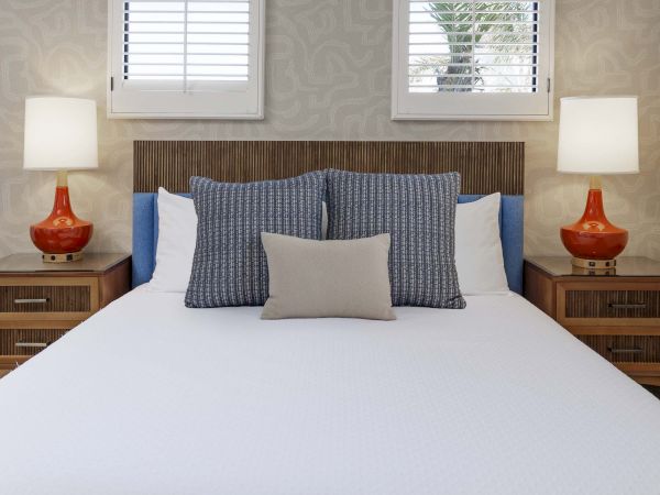 A styled bedroom with a neatly made bed, blue patterned pillows, wooden headboard, bedside lamps, and two windows with blinds.