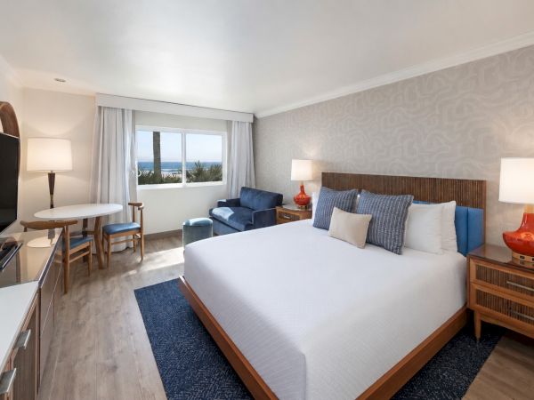 A modern hotel bedroom with a king bed, blue armchairs, lamps, and a view of the ocean through curtains.