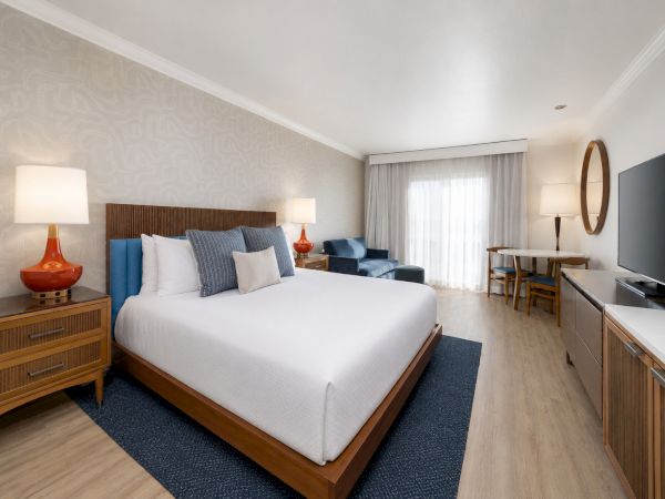 A modern hotel bedroom with a large bed, blue accents, bedside lamps, a seating area, and a window with curtains.