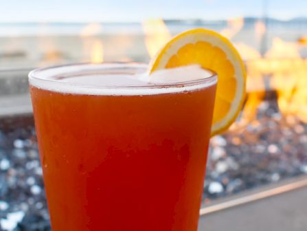 A bright amber cocktail with a lemon wheel, set on a glass-topped table near a beach fire pit, flanked by orange flames in the background.