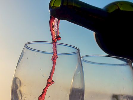 A dark bottle pours red wine into two clear glasses in sunlight, creating a dramatic stream against the blue sky.