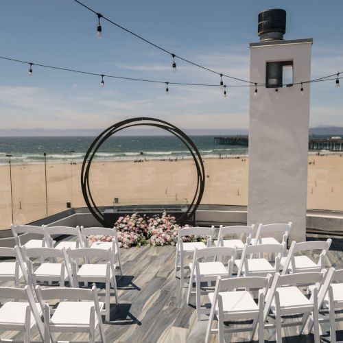 A beach wedding ceremony setup with white chairs, floral decor, hanging string lights, and an ocean view.
