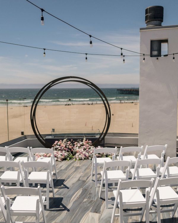A beach wedding ceremony setup with white chairs, floral decor, hanging string lights, and an ocean view.