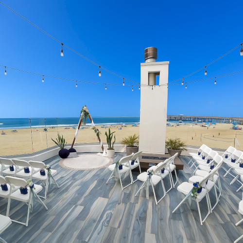 A seaside wedding setup on a wooden deck with white chairs facing the ocean, string lights overhead, and a small tower in the center.
