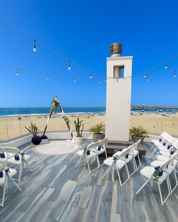 A seaside wedding setup on a wooden deck with white chairs facing the ocean, string lights overhead, and a small tower in the center.