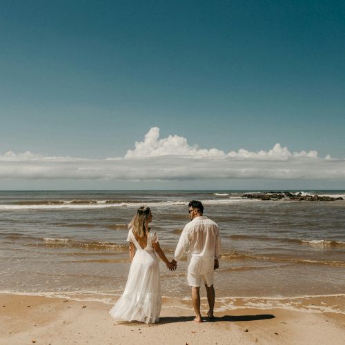 A couple in wedding attire walk hand-in-hand along a quiet beach, waves kiss the shore under a clear, sunny sky.