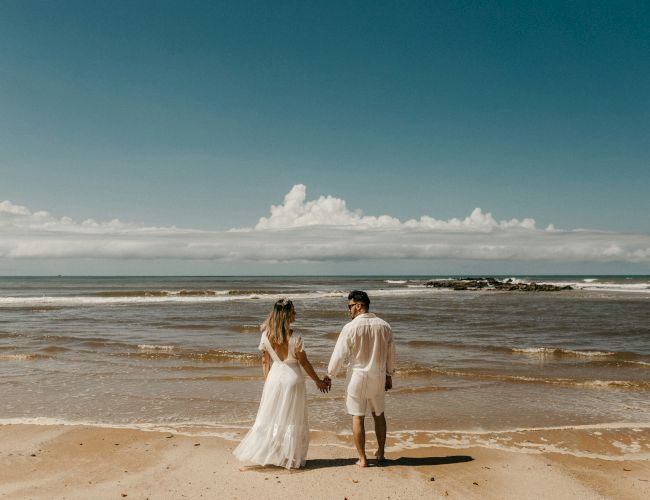 A couple in wedding attire walk hand-in-hand along a quiet beach, waves kiss the shore under a clear, sunny sky.