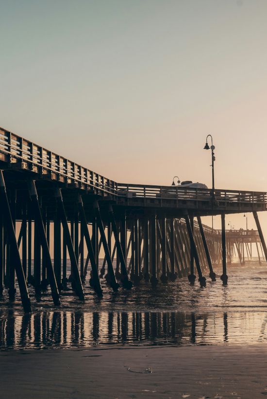 A wooden pier extends over calm sea at sunset, with stilts dipping into the water and a warm, hazy sky in the distance.
