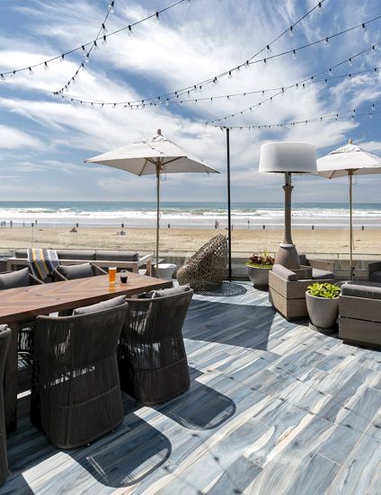 A stylish beachfront patio with wicker seating, a long wooden table, sun umbrellas, string lights, and a view of the sandy beach and ocean under a blue sky.
