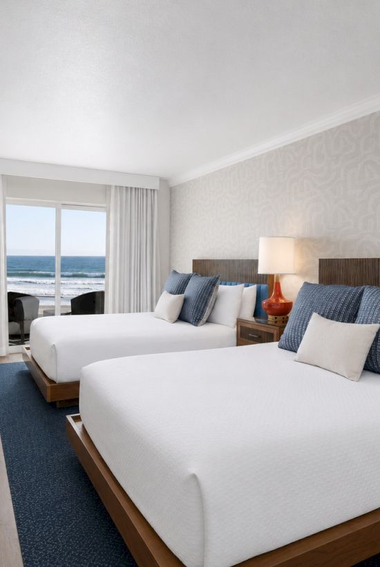 A modern hotel bedroom suite with an ocean view, beds with pillows, bedside lamps, a TV, and a small dining table.