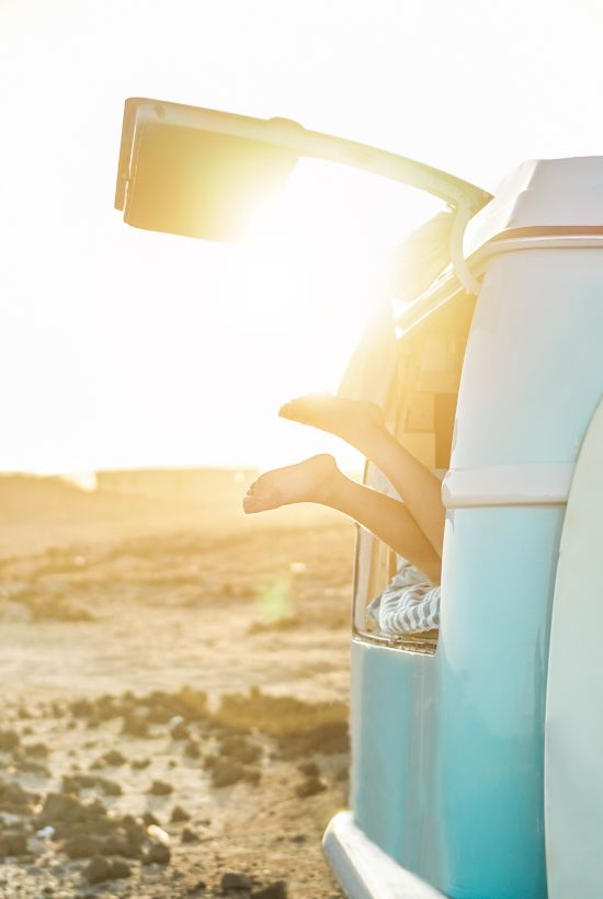 A vintage blue van with a surfboard on the side, sun blazing behind; beachy, retro vibes as the tailgate opens by the shore.
