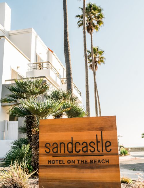 A wooden sign reads Sandcastle Hotel on the Beach beside palm trees and a white beachside building.