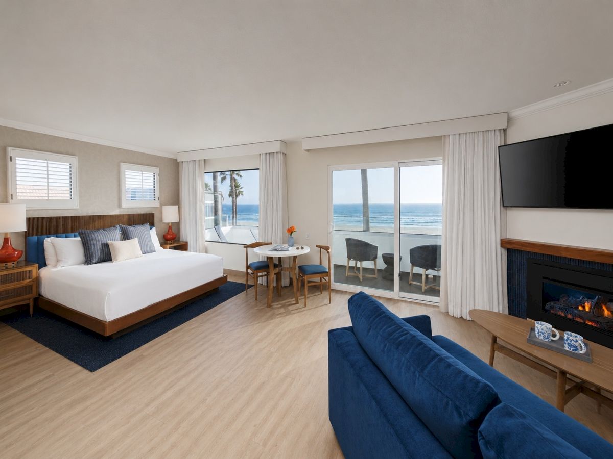A modern ocean-view living bedroom with a king bed, blue sofa, coffee table, TV, and a fireplace, overlooking palm trees.