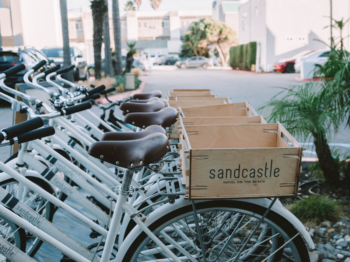 A row of white bikes with brown seats and a cardboard box labeled &ldquo;sandcastle&rdquo; on the front, parked along a sunny street.