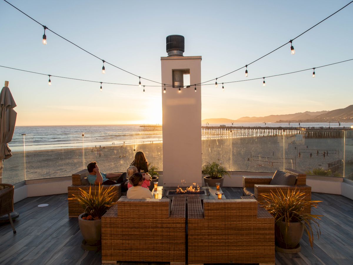 A sunny rooftop lounge by the sea with string lights, a small bell tower, wicker furniture, and guests enjoying the ocean view at sunset.