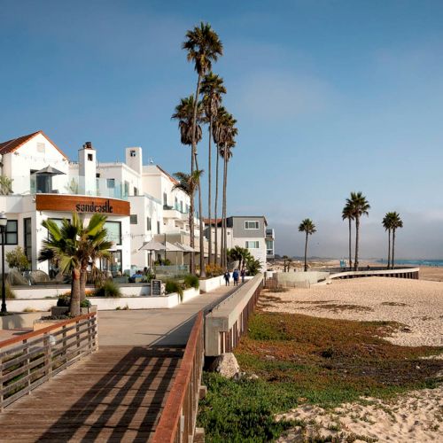 A sunny coastal town with white-washed buildings, a clear blue sea, and people strolling along a wooden boardwalk by the beach.