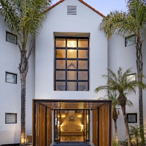 A modern white building entrance with a glass door, wooden frame walkway, and palm trees framing the courtyard in warm light.