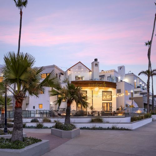 A sunny courtyard at dusk with tall palm trees and string lights, featuring a &ldquo;sandcastle&rdquo; sign on a white hotel.