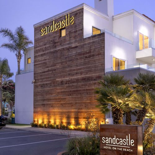 A modern hotel exterior at dusk with glowing &ldquo;Sandcastle&rdquo; signage and palm landscaping, plus a roadside welcome sign.