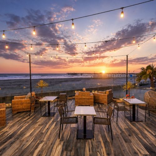 A beachside patio at sunset with string lights, wicker tables, and chairs facing the ocean.