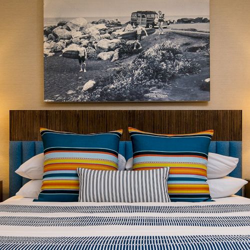 A hotel bedroom with two lamps, striped pillows, a wooden headboard, and a wall photo of a bus by the rocks.