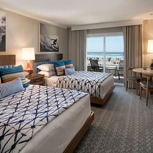 Modern hotel suite with two beds and blue-striped decor, lamps, and a window showing the ocean view.