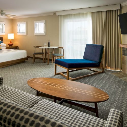 A modern hotel suite with a bed, lounge chair by a fireplace, TV, and coffee table.