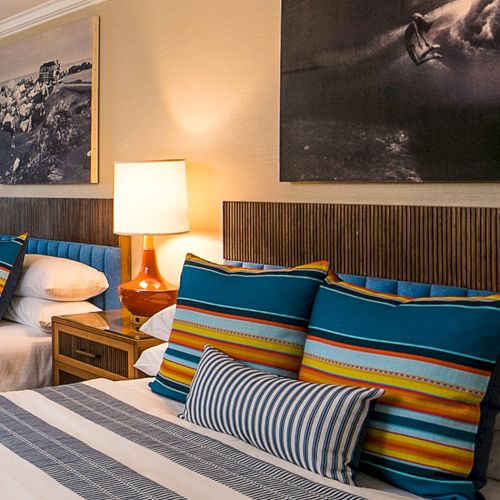 A cozy hotel bedroom with two beds, colorful striped pillows, bedside lamps, and large wall photos.
