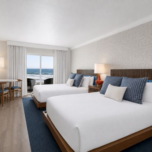 Bright hotel room with beds, blue pillows, lamps, TV, and a large window showing the ocean.