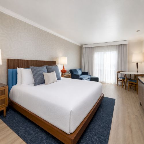 Modern hotel bedroom with a large bed, blue throw pillows, lamps, a sofa, curtains, and a TV console.