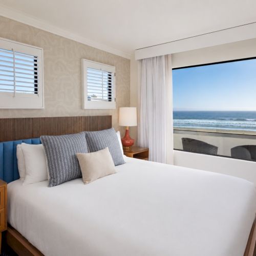 A bright ocean-view bedroom with a neatly made bed, side lamps, patterned walls, and open curtains.