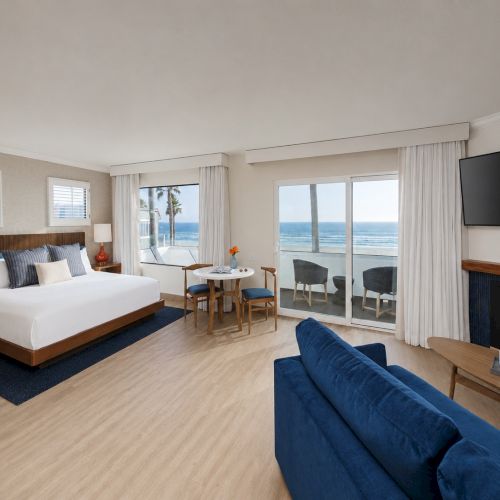 A bright hotel suite with a bed, blue sofa, TV, fireplace, and floor-to-ceiling windows showing the ocean and palm trees.