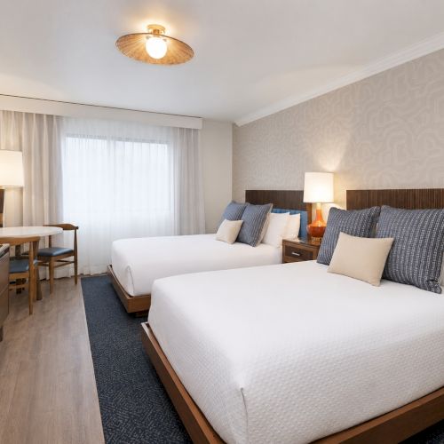 A modern hotel room with two beds, lamps, curtains, patterned wallpaper, and a TV stand.
