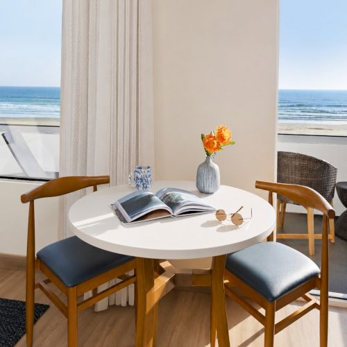 A bright beachfront dining nook with a round table, blue chairs, an open book, flowers, and an ocean view through windows.