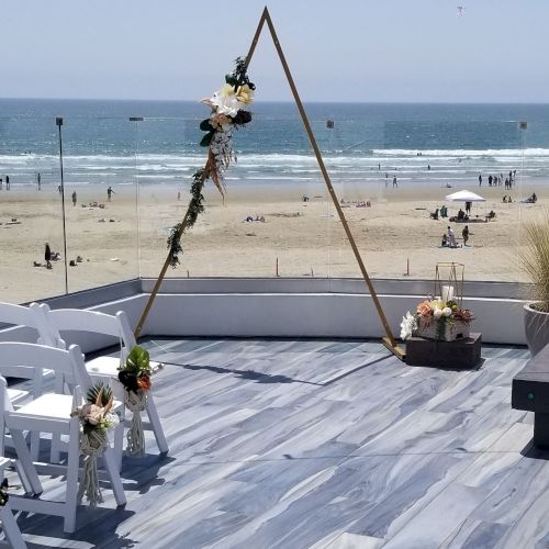 Beachside ceremony setup with white chairs, a floral arch, and ocean waves under an overcast sky.