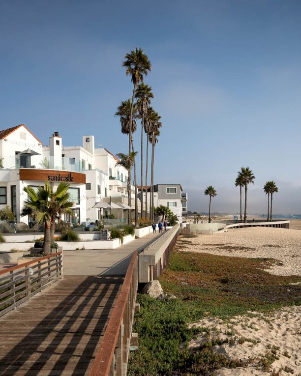 A sunny beachside street with white Mediterranean buildings, palm trees, a boardwalk, and the sandy shore meeting a calm ocean, inviting and peaceful.