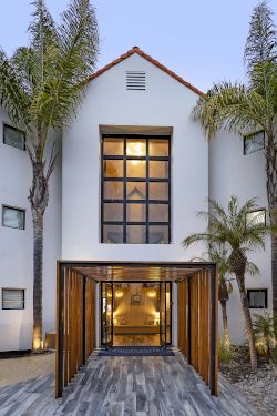 A modern white building entrance with wooden gates, a lit interior, and palm trees framing a small courtyard walkway.