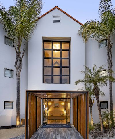 A modern white building entrance with wooden gates, a lit interior, and palm trees framing a small courtyard walkway.
