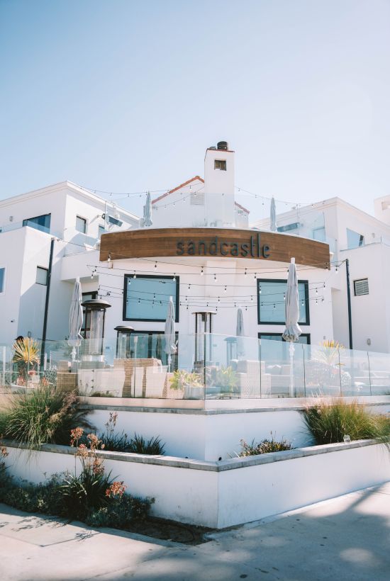 A stylish white storefront with a wooden sign reading &ldquo;sandcastle,&rdquo; glass railings, and coastal landscaping under a bright blue sky.