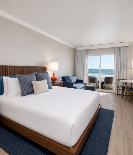 A bright hotel bedroom with a large bed, lamps, seating, and a TV, with curtains opening to an ocean view.