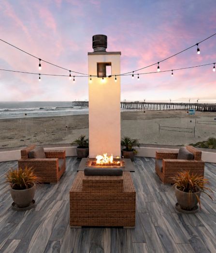 A beachfront patio at dusk with a central fire pit, wicker seating, and hanging string lights overlooking the ocean.