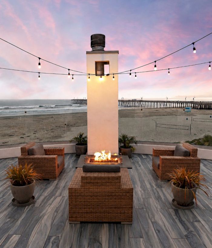 A beachfront patio at dusk with a central fire pit, wicker seating, and hanging string lights overlooking the ocean.