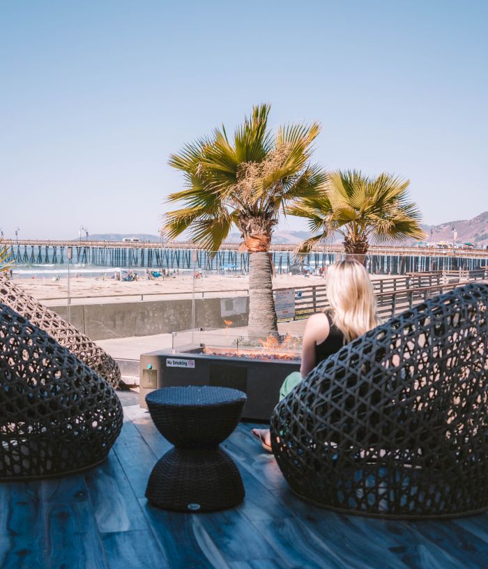 A woman sits on a blue-tiled deck beside two woven basket chairs, facing a sea view with palm trees and a distant pier under clear skies.