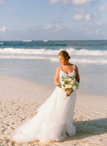 A bride in a white gown stands on a sandy beach, holding a bouquet, with the ocean and waves in the background.