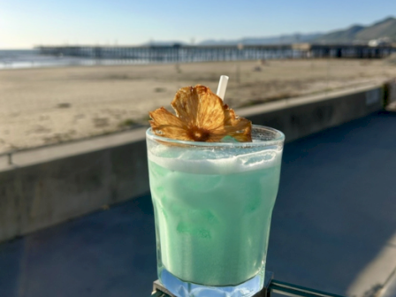 A blue-green cocktail with foam and a crispy garnish sits on a beach promenade under a clear sky.