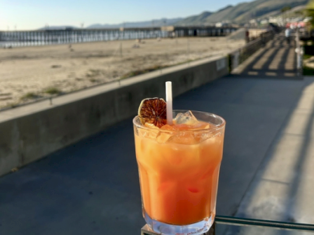 A glass of iced orange drink with a cinnamon stick sits on a seaside boardwalk overlooking sand and distant hills.