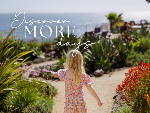 A little girl in a floral dress walks along a coastal garden path toward the ocean, with &ldquo;Discover MORE days&rdquo; overlaid.