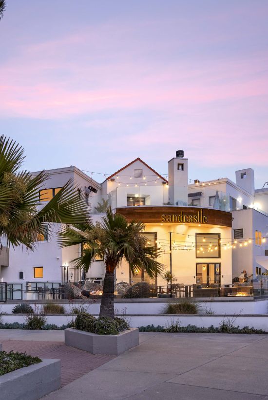 A seaside hotel terrace labeled &ldquo;sandcastle&rdquo; with warm lights, palm trees, and a pink sunset sky.