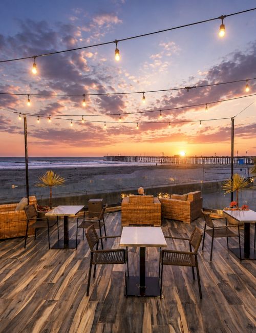 A beachside patio at sunset with string lights, tables, and chairs overlooking the ocean and pier.