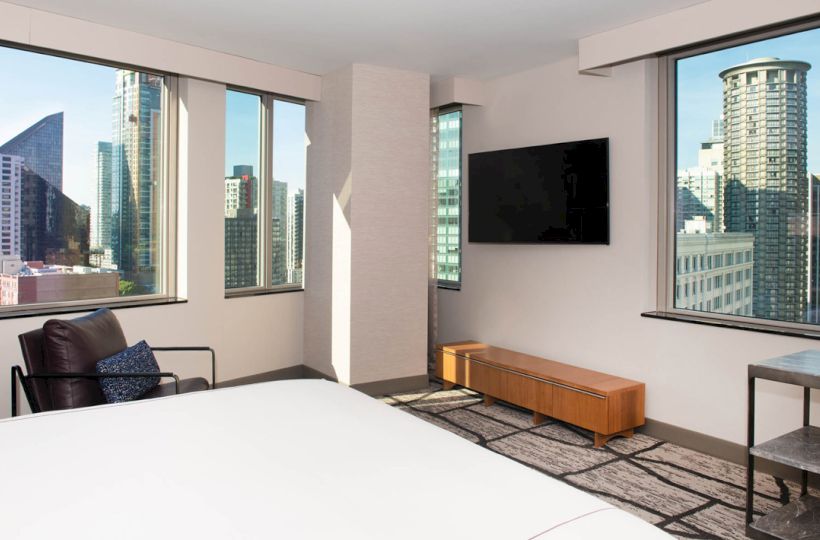 A modern hotel bedroom with a bed, TV console, and large windows showing a city skyline.